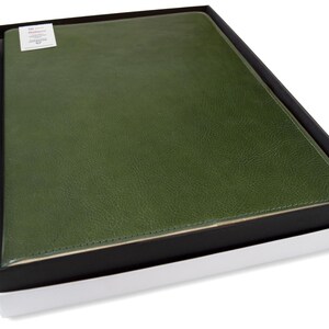 Puede incluir: Un portafolio de cuero verde en una caja negra y blanca. El portafolio tiene una superficie texturizada y un borde cosido. Una peque&ntilde;a etiqueta blanca es visible en la esquina superior izquierda.