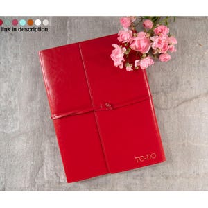 Peut inclure: Un journal relié en cuir rouge avec une fermeture en ruban doré et le mot "TO-DO" en relief sur la couverture. Le journal est posé sur une surface grise avec des fleurs roses.