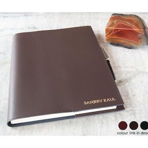 May include: A brown leather-bound journal with the name "SANJEEV KAUL" embossed in gold lettering. The journal is closed and lying on a light brown surface.