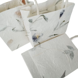 K&ouml;nnte beinhalten: Drei wei&szlig;e Geschenkt&uuml;ten aus Papier mit Blumendesigns und Kordelgriffen. Die Taschen sind aus Recyclingpapier hergestellt und haben ein nat&uuml;rliches, rustikales Aussehen.