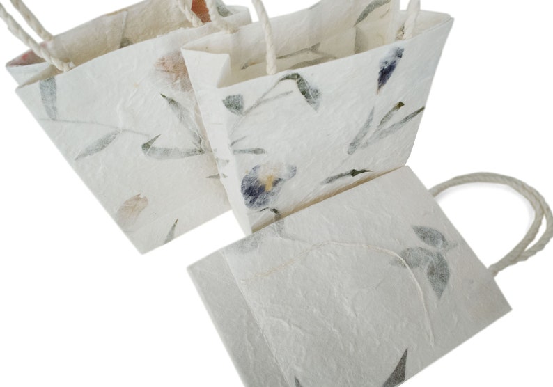 K&ouml;nnte beinhalten: Drei wei&szlig;e Geschenkt&uuml;ten aus Papier mit Blumendesigns und Kordelgriffen. Die Taschen sind aus Recyclingpapier hergestellt und haben ein nat&uuml;rliches, rustikales Aussehen.