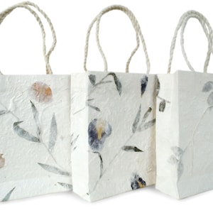 K&ouml;nnte beinhalten: Drei wei&szlig;e Papier-Geschenkbeutel mit Blumendesigns und nat&uuml;rlichen Kordelgriffen. Die Beutel haben eine strukturierte Oberfl&auml;che und zeigen gepresste Blumen und Bl&auml;tter.