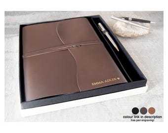 Könnte beinhalten: Ein braunes Lederjournal mit passendem Stift, präsentiert in einer schwarz-weißen Geschenkbox. Das Journal ist mit dem Namen "EMMA ADLER" und einem Herzsymbol personalisiert. Zwei zusätzliche Stifte liegen auf einem dekorativen Stein.