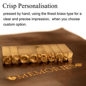 May include: Close-up of a set of brass letter stamps with various designs, including an infinity symbol, a heart, an anchor, and a tree. The stamps are arranged in a row on a brown leather surface with the word "MEMORIES" embossed in gold lettering.