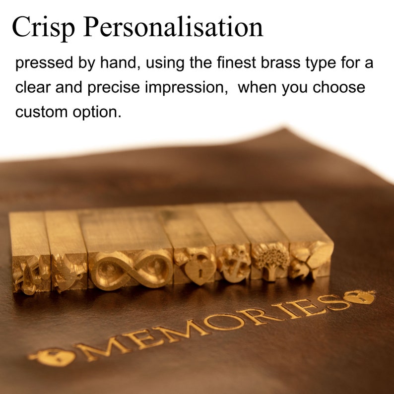 May include: Close-up of a set of brass letter stamps with various designs, including an infinity symbol, a heart, an anchor, and a tree. The stamps are arranged in a row on a brown leather surface with the word "MEMORIES" embossed in gold lettering.