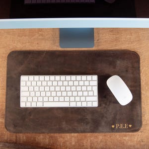 May include: A brown leather desk mat with a white keyboard and mouse. The mat has rounded corners and the initials "P.E.E." with a heart symbol are engraved on the bottom right corner. The keyboard is a standard size.