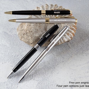 three pens with personalized names engraved on them, placed on a rock or seashell.