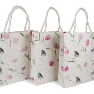 K&ouml;nnte beinhalten: Drei wei&szlig;e Papiert&uuml;ten mit rosa und blauen Blumendesigns und wei&szlig;en Kordelgriffen.