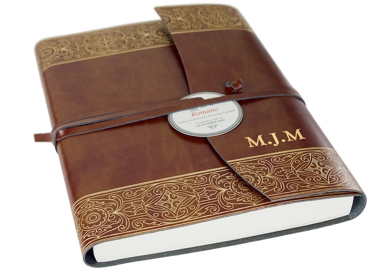 May include: A brown leather journal with gold embossed detailing and a leather strap closure. The journal has a label that reads "Romano" and is personalized with the initials "M.J.M."