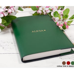 Könnte beinhalten: Ein dunkelgrünes Notizbuch mit dem Namen "ALICIA" in goldfarbenen Buchstaben und einem Herzsymbol. Das Notizbuch ist geöffnet und zeigt weiße Seiten. Rosa und weiße Blumen sind im Hintergrund. Ein Stift steckt im Buchrücken.