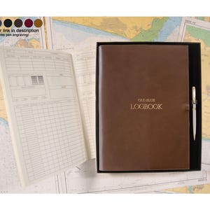 May include: A brown logbook with the text OLD BLUE LOGBOOK embossed in gold on the cover, presented in a black box with a silver pen. The open logbook shows white pages with printed grids for data entry, including columns for date and time.