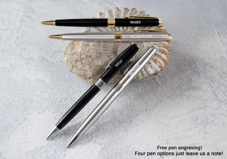 three pens with personalized names engraved on them, placed on a rock or seashell.