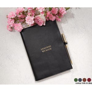 May include: A black leather journal with the gold text "MEMORIES WE MADE" is displayed with a gold pen and pink flowers. The journal is closed and has a smooth texture. The background is a light gray.