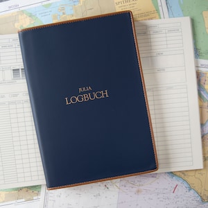 May include: A blue leather-bound logbook with the title "JULIA LOGBUCH" embossed in gold lettering. The logbook is lying on top of nautical charts.