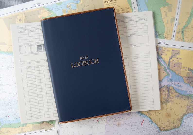 May include: A blue leather-bound logbook with the title "JULIA LOGBUCH" embossed in gold lettering. The logbook is lying on top of nautical charts.