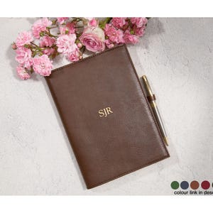 May include: A brown leather journal with the initials "SJR" in gold lettering. A gold pen rests on the right side of the journal. Pink flowers are in the upper left corner. The journal is on a white surface.