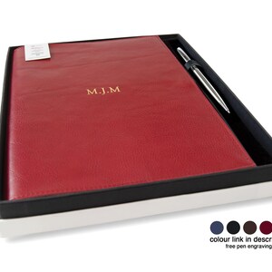 May include: A red rectangular notebook with the gold embossed initials M.J.M. on the cover, presented in a black and white gift box alongside a silver metal pen. The set includes a small white tag and a color selection guide at the bottom.