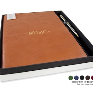 May include: A tan leather-textured notebook with the word MUSIC embossed in gold on the cover, presented in a black and white gift box alongside a silver-toned pen. The set includes a small white tag on the notebook and a color selection guide.
