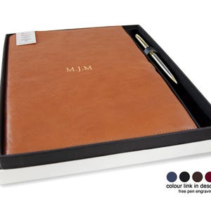 May include: A tan leather-like notebook with the gold embossed initials M.J.M. centered on the cover, presented in a black and white gift box alongside a silver and gold pen. The box includes a color chart with five circular swatches.