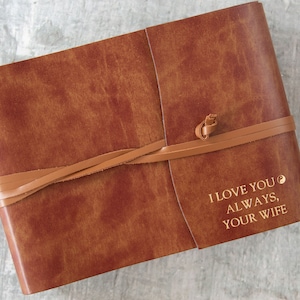 Puede incluir: Un diario encuadernado en cuero marr&oacute;n con cierre de correa de cuero. El diario tiene el texto "I love you always, your wife" grabado en oro en la portada.