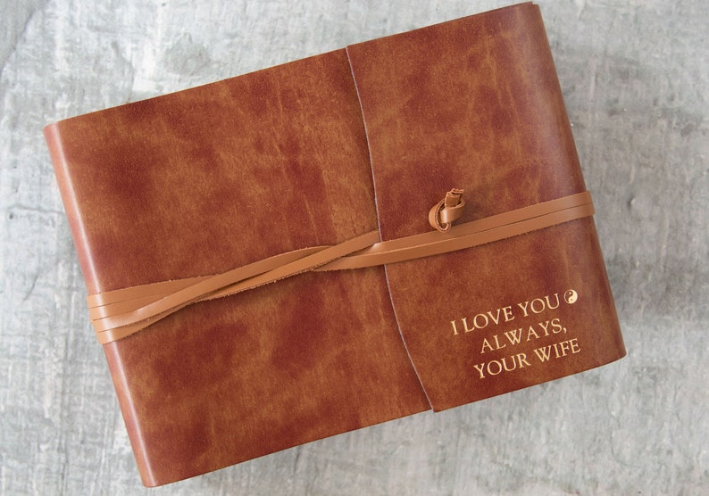 Puede incluir: Un diario encuadernado en cuero marr&oacute;n con cierre de correa de cuero. El diario tiene el texto "I love you always, your wife" grabado en oro en la portada.