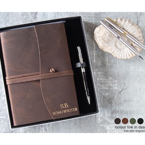 May include: A brown leather journal with a matching pen, presented in a black gift box. The journal is personalized with initials and the word "SONGWRITER". Two silver pens rest on a decorative stone. A thoughtful gift.