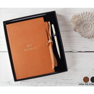May include: A tan leather-bound journal with a wrap-around tie closure, embossed with the text SES VOLUME III, presented in a black gift box alongside a silver-toned ballpoint pen. The journal features a smooth finish and a matching leather tie.