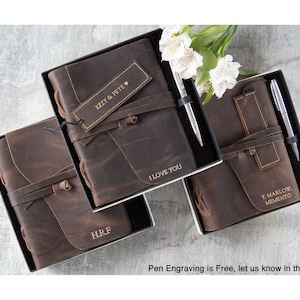 May include: Brown leather journals in black boxes, each with a pen. The journals have a strap closure and personalized text: "I LOVE YOU", "EZZY & PETE", "H.R.F", and "T. MARLOW, MEMENTO". White flowers are in the background.