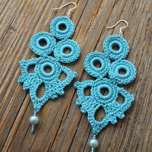 Puede incluir: Un par de pendientes de ganchillo de color azul turquesa. Los pendientes están hechos de varios círculos y tienen una perla en la parte inferior de cada pendiente.