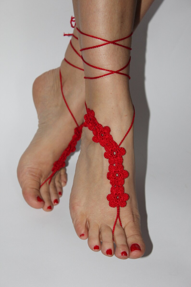 May include: Red crocheted barefoot sandals with flower accents. The sandals are tied around the ankles with red string.