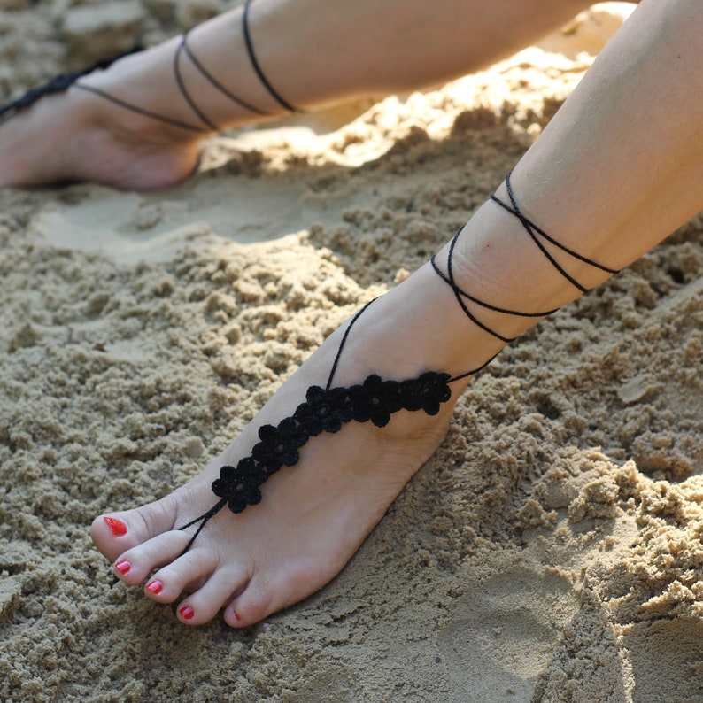 May include: Black crocheted barefoot sandals with flower accents. The sandals are tied around the ankle and foot.