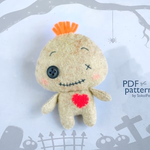 May include: A felt voodoo doll with a stitched-on heart, a button eye, and a cross stitched on its forehead. The doll has a bright orange hair tuft and is smiling. The background is a light gray with a silhouette of a graveyard and a spider.