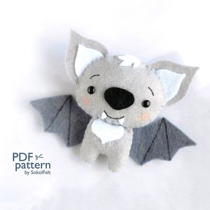 Op de afbeelding: Een grijze vleermuis van vilt met witte oren en een witte borst. De vleermuis heeft zwarte ogen en een witte glimlach met twee hoektanden. De vleermuis heeft grijze viltvleugels.