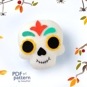 May include: A white felt skull with a colorful Day of the Dead design. The skull has black eyes, a yellow flower design, and a red and green floral pattern on the forehead. The skull is surrounded by a white background with a watercolor design of branches and leaves.