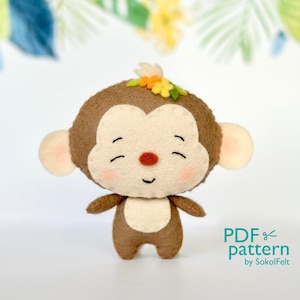 Può includere: Un giocattolo di scimmia di feltro marrone con un viso sorridente e una corona di fiori. La scimmia è in piedi su una superficie bianca. Modello PDF di SokolFelt.