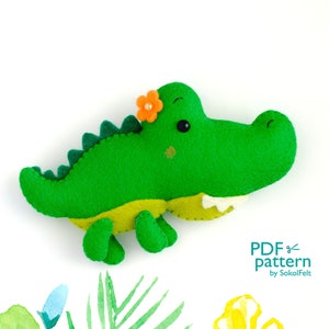 Könnte beinhalten: Grünes Filz-Alligator-Spielzeug mit einer orangefarbenen Blume auf dem Kopf. Das Spielzeug hat einen gelben Bauch und weiße Zähne. Der Text "PDF pattern by SokolFelt" befindet sich in der unteren rechten Ecke des Bildes.