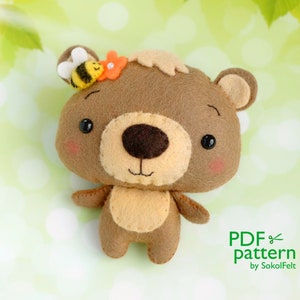 May include: A brown felt bear with a bee and flower on its head. The bear has a cute face with black eyes and a brown nose. The bear is standing on a green and white background. PDF pattern by SokolFelt.