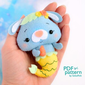 Op de afbeelding: Een blauwe vilt konijn zeemeermin met een gele staart en een groene bloemenkroon. Het konijn houdt een wortel in zijn hand. Het konijn zit in een hand. De tekst "PDF pattern by SokolFelt" staat in de rechterbenedenhoek van de afbeelding.