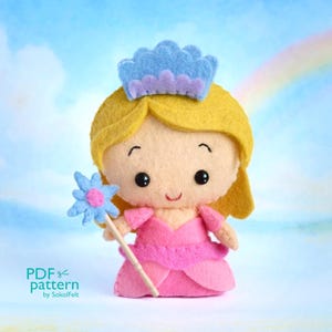 May include: A handmade felt doll of a princess wearing a pink dress and a blue crown. The doll has blonde hair and is holding a blue flower wand. The background is a blue sky with a rainbow. The text "PDF pattern by SokolFelt" is in the lower left corner.