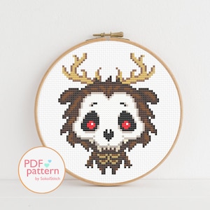 Puede incluir: Patrón de punto de cruz de un lindo ciervo de dibujos animados con cara de calavera y ojos rojos. El ciervo es marrón con cuernos dorados. El patrón está sobre un fondo blanco.