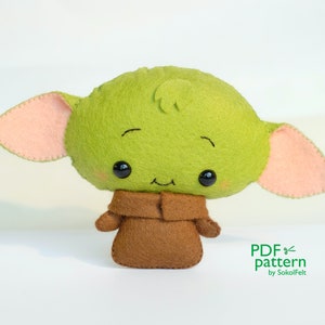 May include: A green felt plush toy with large pink ears and a brown felt robe. The toy has a cute, cartoon-like face with black eyes and a small smile. The text "PDF pattern by SakalFelt" is visible in the bottom right corner of the image.