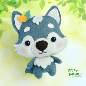 May include: A gray felt wolf with a white belly and a yellow flower on its head. The wolf has black eyes and a pink nose. The background is a blurry green and yellow.