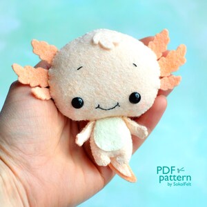 May include: A plush axolotl toy with orange feathery gills, black eyes, and a white belly. The toy is made of felt and is being held in a hand. The text "PDF pattern by SokolFelt" is visible in the bottom right corner of the image.