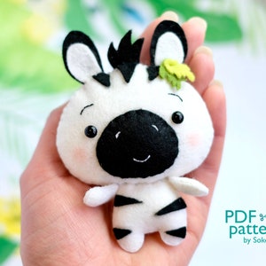 May include: A white and black felt zebra plush toy with a green leaf on its head. The zebra has a black nose and black eyes. The toy is being held in a person's hand.