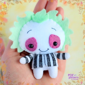 May include: A white felt plush toy with black and white striped clothing, green hair, and pink eyes. The toy is smiling and has a stitched-on mouth. The toy is being held in a hand. The text "PDF pattern by SokolFelt" is in the bottom right corner of the image.