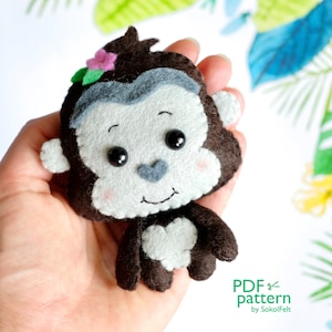 May include: A handmade felt monkey with a smiling face, dark brown fur, and a light gray face and ears. The monkey has a small pink flower and green leaves on its head. The text "PDF pattern by SokolFelt" is in the lower right corner.