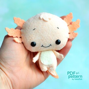 May include: A plush axolotl toy with a light pink body, white belly, and orange feathery gills. The toy has black eyes and a small smile. The toy is being held in a hand. The text "PDF pattern by SokolFelt" is in the lower right corner of the image.