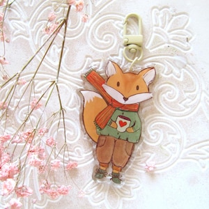 Könnte beinhalten: Ein fuchsförmiger Schlüsselanhänger mit Cartoon-Design. Der Fuchs trägt einen grünen Pullover, eine braune Hose und einen roten Schal. Er hält eine Tasse mit einem Herz. Der Schlüsselanhänger hat einen goldfarbenen Verschluss.