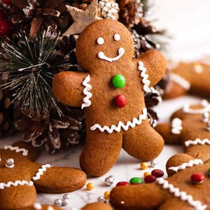 May include: A gingerbread man biscuit decorated with white icing, featuring a smiling face and zig-zag edges. It has green and red candy buttons. Other gingerbread biscuits and festive decorations are in the background.