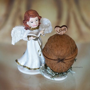 May include: A figurine of an angel in a white gown with gold trim, holding a book. Beside the angel is a walnut with a wooden heart on top that says "Love". The angel has red hair and white wings.